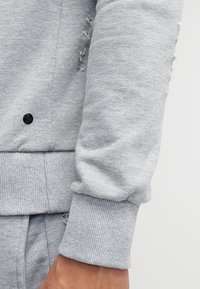 Primo piano del braccio di una persona che indossa una felpa con polsino a coste di colore grigio chiaro, con piccoli fori intenzionalmente consumati vicino al gomito.