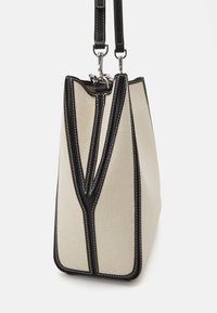 Sac cabas en toile beige avec bordure en cuir noir et coutures blanches, vue de côté avec deux sangles en cuir noir détachables et fermoirs argentés.