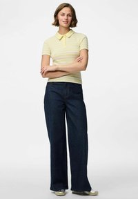Rumena in bela črtasta polo majica z ovratnikom, v kombinaciji s temno modrimi širokimi hlačami. Model stoji s križanimi rokami.