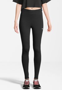 Leggings negros de material elástico, con una cinturilla alta y un pequeño logo en el lado izquierdo, combinados con zapatillas negras y rosas.