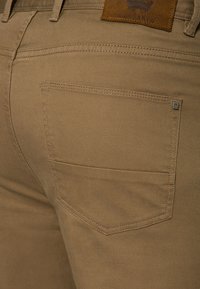 Beżowe bawełniane spodnie o gładkiej fakturze, z tylną kieszenią o delikatnym zaokrągleniu i brązową skórzaną łatą powyżej.