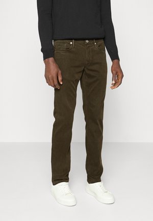 Man wearing dark olive green corduroy pants, black long-sleeve top, and white sneakers, standing against a plain light background.