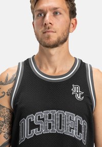 Čierny priehľadný basketbalový dres s šedými pruhmi, výrazným logom "DCSHOECO" a malým vyšívaným logom na hrudi.