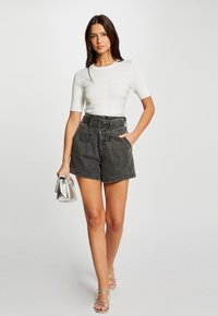 Witte geribbelde top met korte mouwen en hoge zwarte denim shorts met een riem. Het model houdt een kleine zilveren handtas vast en draagt sandalen met bandjes.