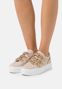 Beżowe zamszowe sneakersy z złotymi ćwiekami, białą podeszwą, niskim profilem i gładką teksturą. Odpowiednie na codzienne wyjścia.