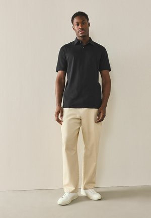 Mann steht vor einer schlichten Wand, trägt ein schwarzes Poloshirt mit kurzen Ärmeln, beige Hosen und weiße Turnschuhe, blickt nach vorne und hat eine entspannte Haltung.