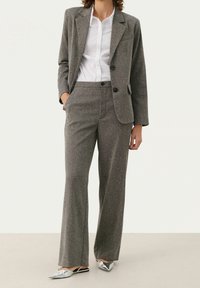 Femme portant un tailleur gris composé d'un blazer cintré et d'un pantalon, avec une chemise blanche boutonnée et des chaussures slingback métalliques à bout pointu.