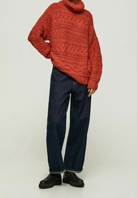 Paksu punainen neulottu pusero tekstuurillisilla kuvioilla ja korkealla kauluksella, yhdistetty löysiin tummiin farkkuihin ja mustiin paksupohjaisiin saappaisiin.