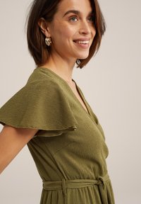 Olívazöld ruha rövid, lebegő ujjakból, texturált anyagból, V-nyakú kivitelben, övvel a deréknál és oldalsó zsebekkel. Arany karika fülbevalók.
