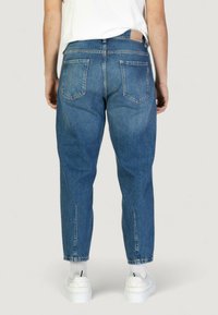 Jeans in denim blu con vestibilità rilassata, dotati di due tasche posteriori e dettagli cuciti. I bordi inferiori presentano delle fessure, abbinati a sneakers bianche.
