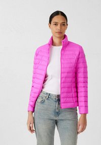 Giacca a vento rosa con design trapuntato, collo alto e apertura frontale con zip. Indossata sopra una camicia bianca e abbinata a jeans di denim chiaro.