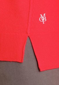Czerwony sweter z prążkowanym wzorem, bocznymi rozporkami oraz małym, białym haftowanym logo na dolnym brzegu. Gładki materiał w intensywnym kolorze.