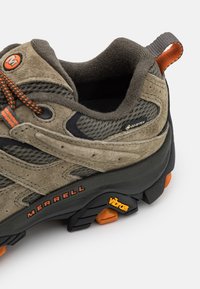 Chaussure de randonnée grise et beige avec des matériaux en daim et en maille, dotée d'accents orange, d'une semelle Vibram visible et d'un système de laçage texturé.