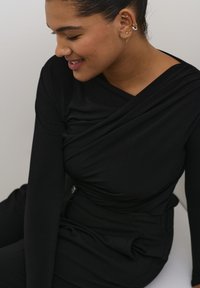 Haut noir à manches longues avec un design drapé, présentant un effet croisé et une texture lisse. Le modèle adopte une pose détendue sur un fond neutre.