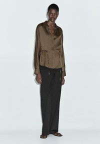 Chemise longue à manches longues en satin marron avec une coupe décontractée, taille ajustable avec cordon, et col pointu, associée à un pantalon large noir.