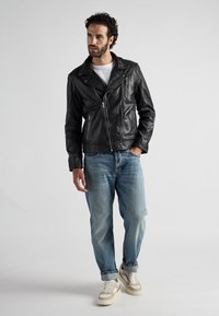 Schwarze Leder-Motorradjacke mit asymmetrischem Reißverschluss, kombiniert mit hellblauen Jeans und beigen Sneakers, präsentiert an einem Modell.
