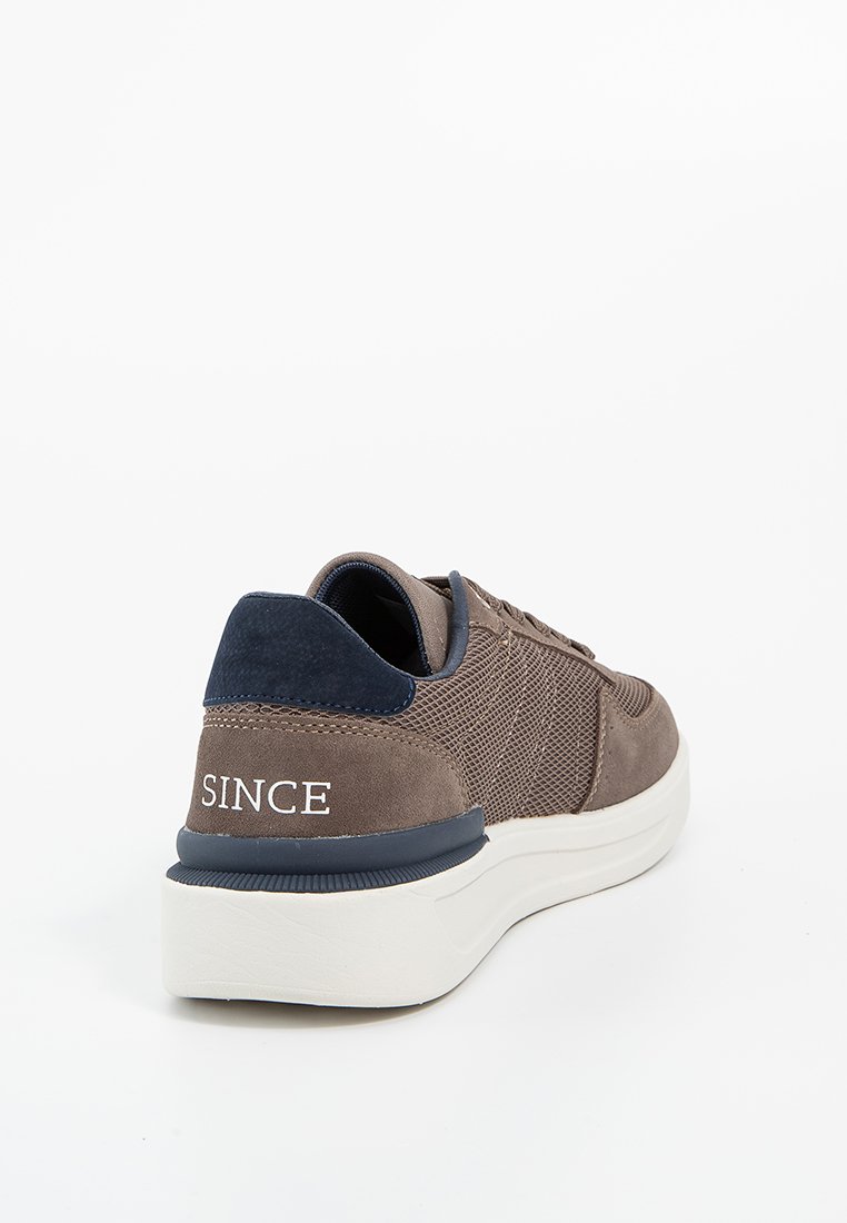 Bruine suede sneaker met marineblauwe accenten, mesh-details en een witte zool. Kenmerkt het woord "SINCE" op de hiel. Laag ontwerp.