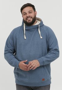 Sudadera azul de tela suave, con cuello alto y cordones, puños acanalados y un pequeño parche del logo en la parte inferior izquierda.