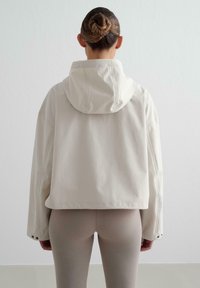 Veste courte couleur crème avec capuche. Présente des manches larges et des accents de boutons-pression. Texture lisse, design minimaliste.
