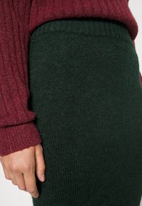 Burgundy stickad tröja med ribbat mönster, ihop med en mörkgrön stickad kjol, som har en slät yta och elastiskt midjeband.