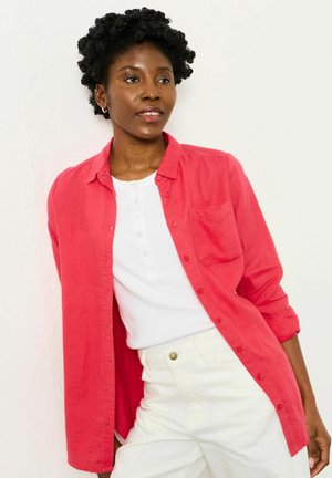 Rosa Hemd mit Knöpfen und Tasche, offen über einem weißen Henley-Oberteil getragen. Das Modell trägt weiße Hosen. Glatter Stoff, lässige Passform.