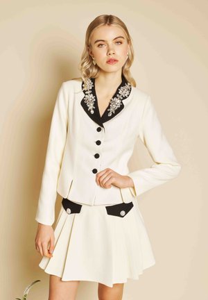 Jeune femme portant un blazer blanc avec un col à bijoux noirs et une jupe plissée assortie avec des accents de poche noirs, debout devant un fond beige.