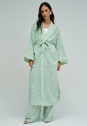 Mujer con una bata larga texturizada verde menta con mangas anchas y pantalones de pierna ancha a juego, atada en la cintura con una faja.