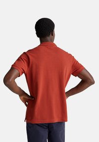 Polo a maniche corte in terracotta, tessuto di cotone. Presenta un colletto classico e fessure laterali all'orlo. La vista posteriore rivela una consistenza liscia.