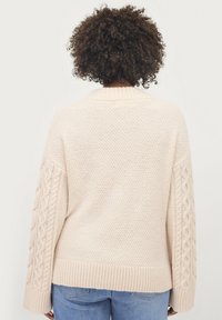 Maglione lavorato a maglia beige chiaro con colletto e orlo a coste, caratterizzato da dettagli a treccia sulle maniche e un corpo testurizzato.