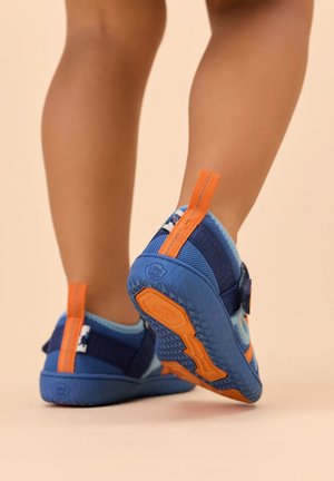 Gambe di un bambino che indossano sneakers blu e arancioni con suole testurizzate, in piedi su uno sfondo beige.
