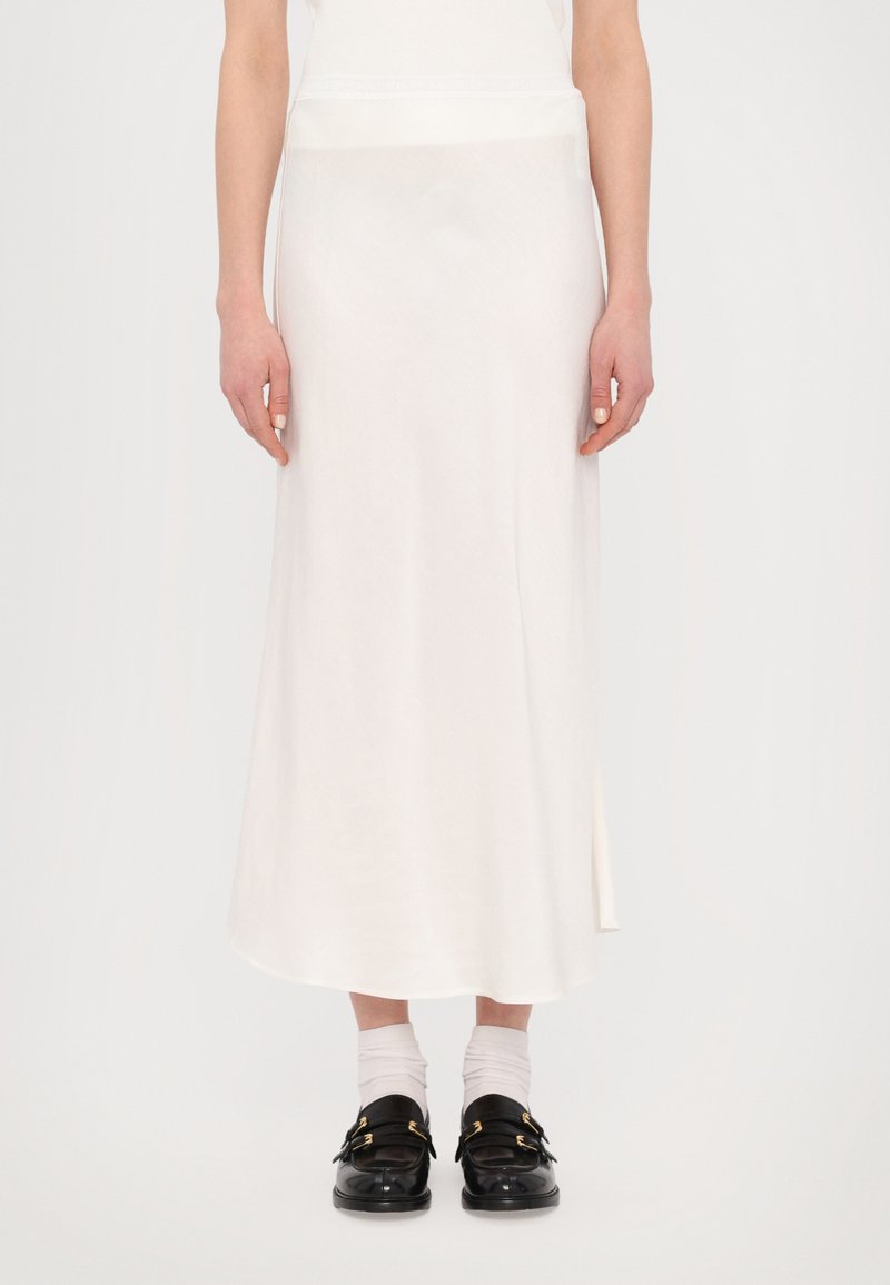 Jupe midi blanche en tissu lisse avec une fente sur le côté et une coupe décontractée. Associée à des chaussures noires et des chaussettes blanches à la cheville.