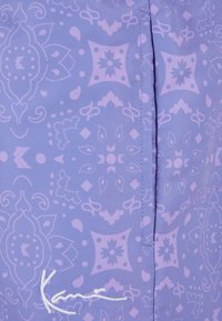 Tissu violet à motifs présentant des designs et formes complexes, avec des accents de lavande clair. Comprend une couture visible et un détail de logo brodé.
