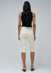 Pantalones capri beige con bolsillos traseros, hechos de tela elástica. Combinados con una camiseta de tirantes negra ajustada, vistos desde la parte de atrás.