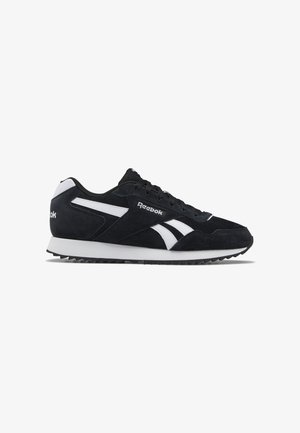 Zapatilla de ante negra con acentos blancos, que presenta un diseño clásico de corte bajo, cierre con cordones y suela de goma texturizada. Marca Reebok visible.