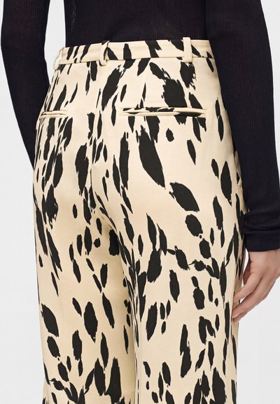 Pantalon beige avec des taches noires irrégulières en motif, poches arrière et passants de ceinture porté avec un haut noir à manches longues.