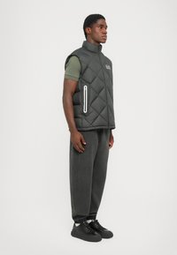Vest cu umplutură neagră, cu quilted, guler înalt, cu buzunare laterale cu fermoar, purtat peste o cămașă verde, asociat cu pantaloni gri și pantofi negri.