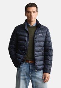 Marinblå vadderad jacka med quiltad design, hög krage och fickor med dragkedja, bärs över en grön tröja och ljusblå jeans.