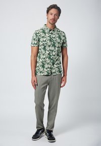 Camicia a maniche corte verde con motivi a foglia bianchi, abbinata a pantaloni grigio chiaro e sneakers nere con dettagli gialli.