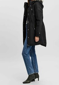 Parka noire à capuche avec doublure en fourrure, taille cintrée, poches avant et fermetures à pression, associée à un jean bleu et des bottines noires.