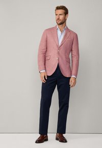 Blazer de lino rosa claro con solapas muescas, cierre de un botón, bolsillo en el pecho; combinado con una camisa azul y pantalones marinos. Zapatos marrones.
