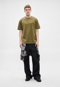 Bombažna majica olivno zelene barve s kratkimi rokavi, kombinirana z ohlapnimi temnimi jeans hlačami in črno-belo karirasto srajco, vezano okoli pasu.