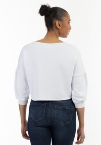 Felpa bianca corta con scollo ampio e maniche a sbuffo, abbinata a jeans aderenti blu scuro, vista da dietro.