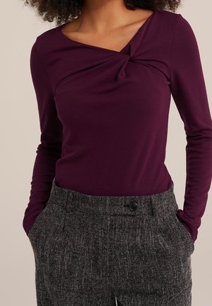 Kobieta w długim rękawie ciemnofioletowego topu z detalem skręconego dekoltu i szarych, teksturowanych, wysokich spodni z rękami w kieszeniach.