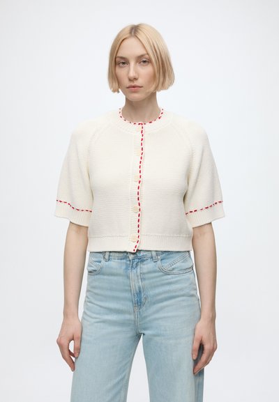Jeune femme aux cheveux blonds courts portant un cardigan blanc en maille à manches courtes avec des coutures rouges et un jean taille haute bleu clair.