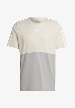 Hombre vistiendo una camiseta Adidas de manga corta en tonos beige y gris con rayas blancas en los hombros y pantalones negros, sobre fondo blanco.