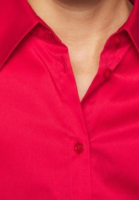 Czerwona koszula zapinana na guziki o gładkiej fakturze, klasycznym kołnierzyku i wyraźnych guzikach. Materiał wydaje się lekki i delikatnie błyszczący.