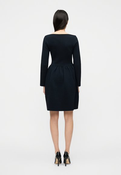 Μαύρο φόρεμα με μακριά μανίκια, εφαρμοστό πάνω μέρος και φουσκωτή φούστα, με διακριτική υφή στο ύφασμα και τετράγωνο ντεκολτέ, φορεμένο με μαύρες ψηλοτάκουνες γόβες.