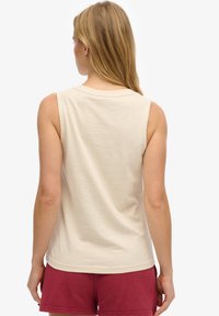 Vrouw met lang blond haar, gekleed in een mouwloos beige topje en bordeauxrode korte broek, staand met de rug naar de camera tegen een witte achtergrond.