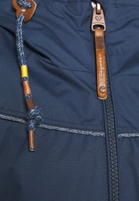 Marinblazer har en texturerad tyg med kontrasterande sömmar, en metalldragkedja och en läderdragflik med ett vävt snöre och färgade accenter.