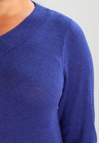 Gros plan d'une personne portant un pull en maille bleu à col en V sur un fond clair uni.
